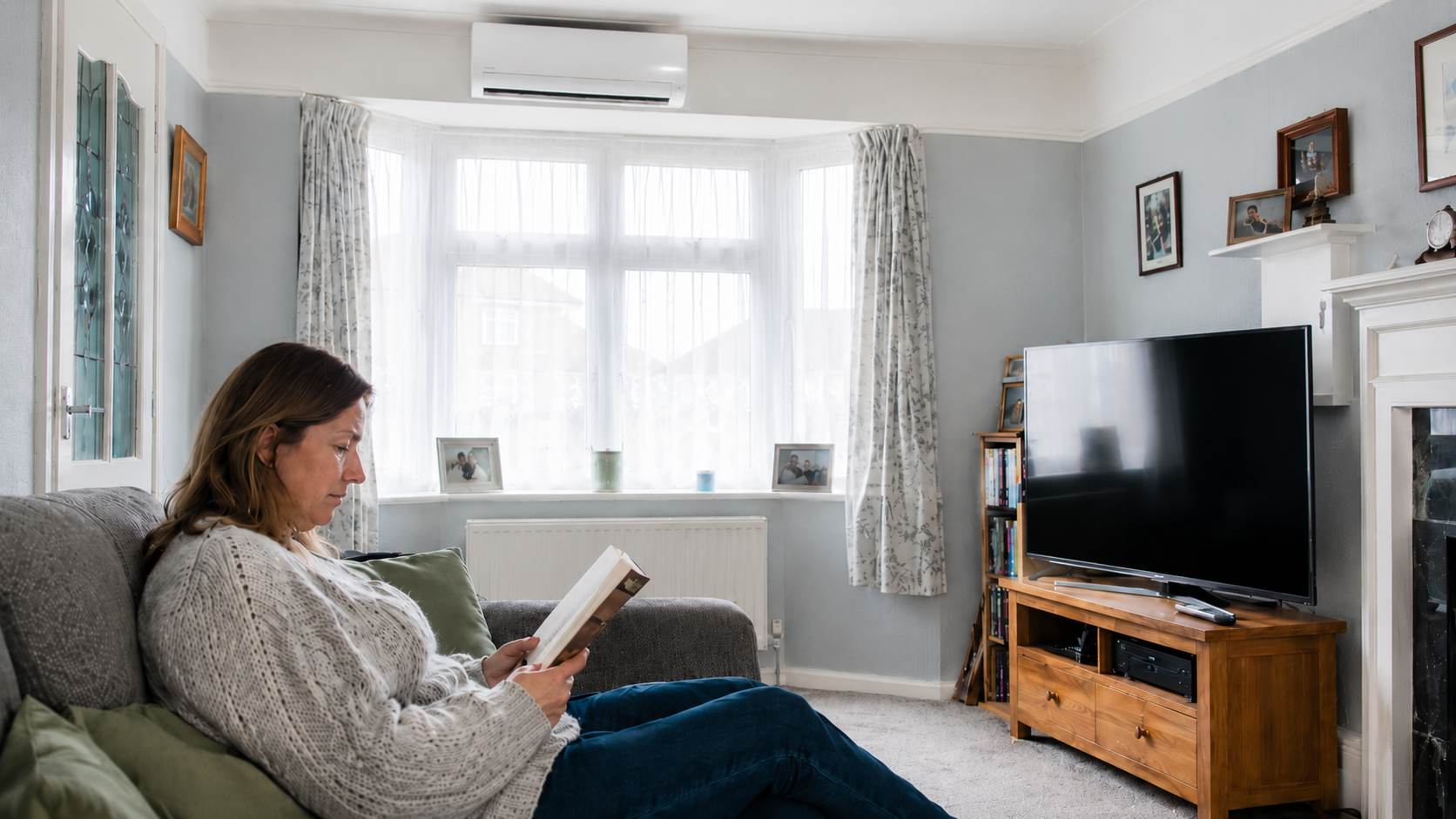 Woman reading on a sofa in a UK living room with a wall-mounted air conditioning unit above the bay window