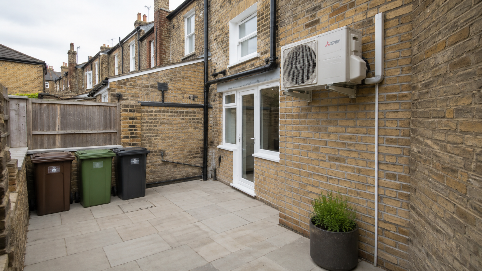 Air conditioning condenser mounted on a brick wall in a narrow enclosed courtyard behind a UK property